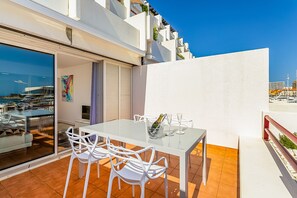 A glass table with white chairs, a bottle of champagne, and two glasses on a balcony with a view of boats and buildings.