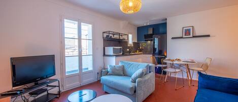 A modern living room with a flat-screen TV, a light blue sofa, a round white table, and a dining area with chairs and a small table.