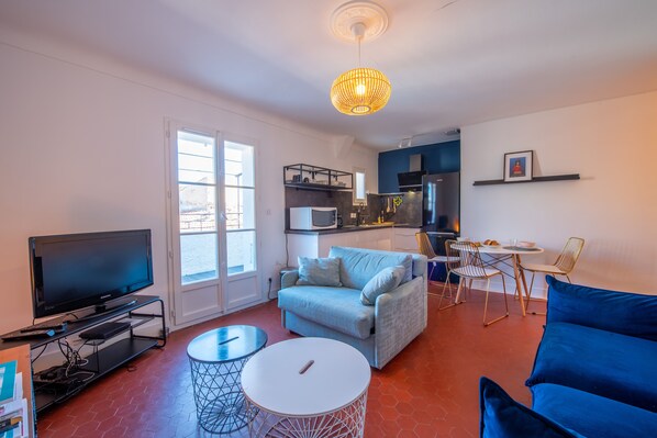 A modern living room with a flat-screen TV, a light blue sofa, a round white table, and a dining area with chairs and a small table.