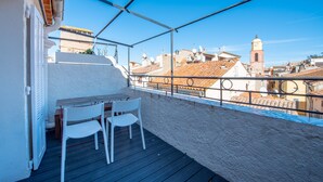 Un balcon avec une table et des chaises, donnant sur un paysage urbain avec des immeubles et une tour de l’horloge.