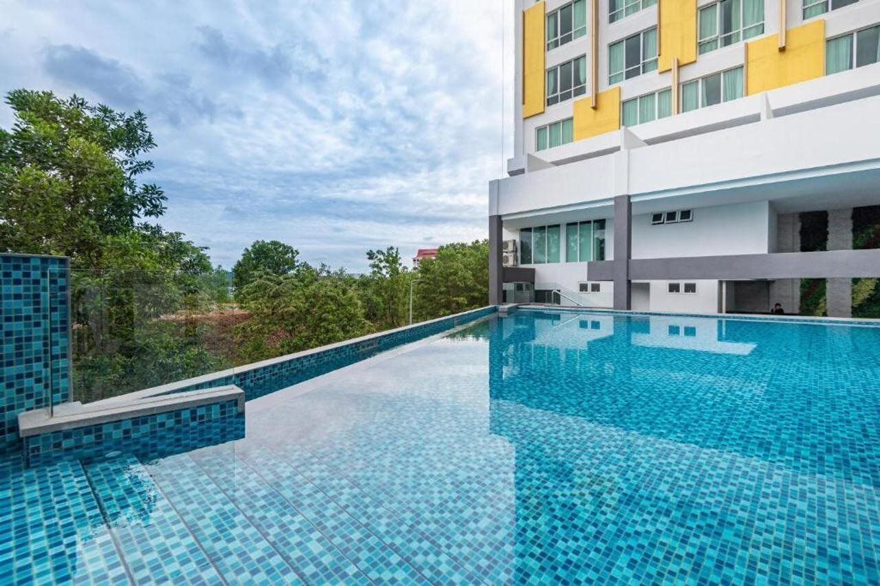 A modern hotel with a rooftop infinity pool, surrounded by lush greenery and a clear sky.