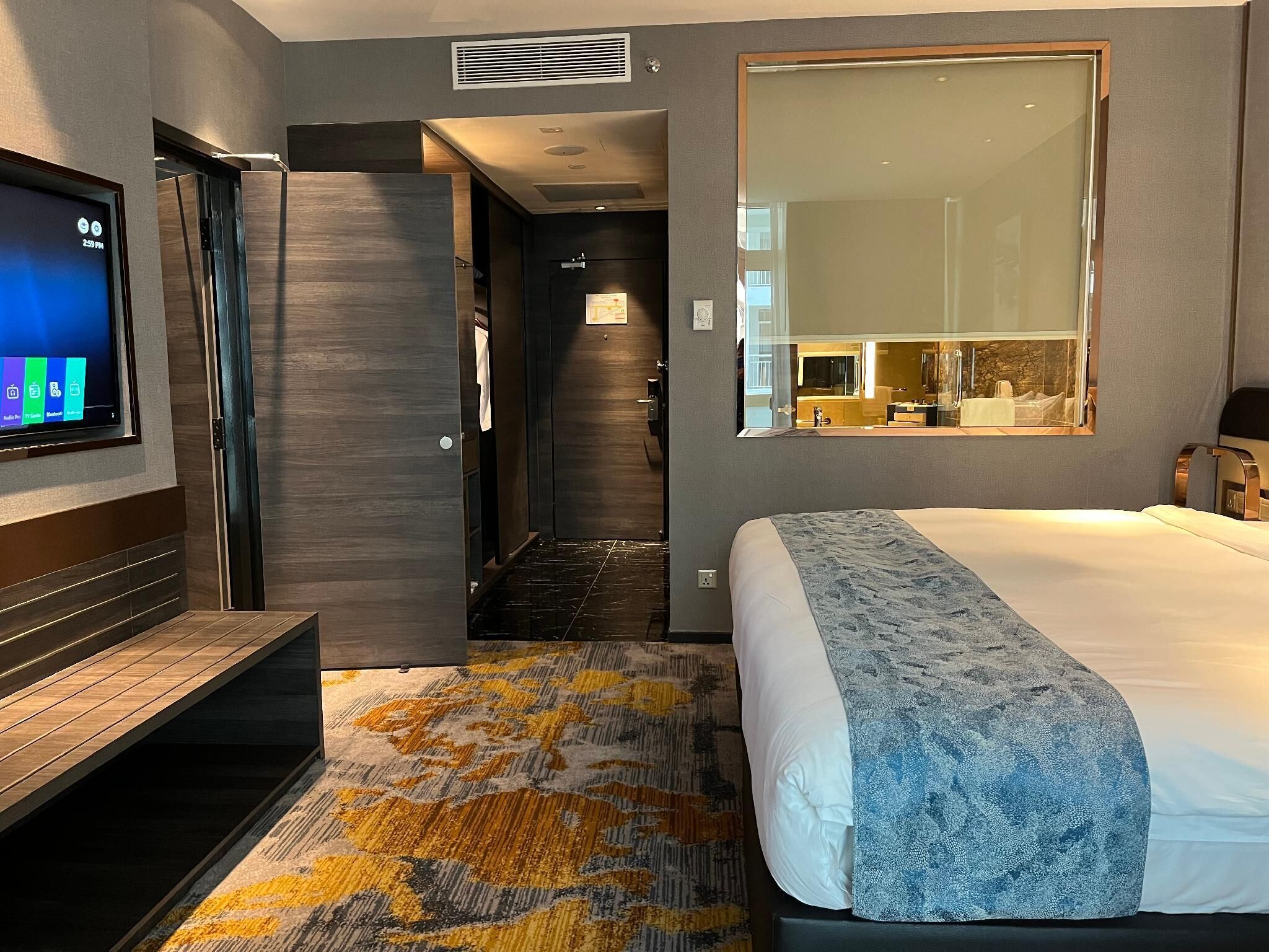 A modern hotel room with a large bed, a TV mounted on the wall, a bathroom with a mirror, and a hallway leading to another room.