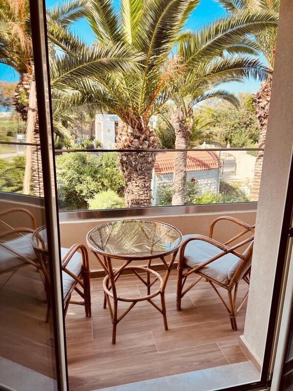 A balcony with wicker chairs and a glass table, overlooking palm trees and a building with a tiled roof. - Lili Hotel (Malevizi)