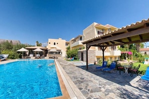Una piscina con sillones de descanso y sombrillas, rodeada de edificios con balcones y un sendero de piedra.