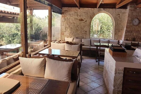 A cozy room with a stone wall, a wooden bench, a table, and a view of greenery outside. - Lili Hotel (Malevizi)