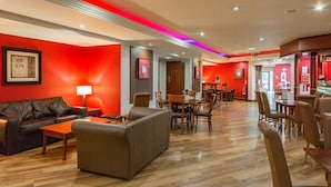 Et hotellobby med rød væg, trægulv, lædersofaer og et spiseområde med borde og stole.