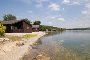 Une cabane au bord d’un lac aux eaux cristallines, avec un chemin de gravier et une terrasse en bois.