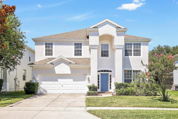 A two-story house with a white exterior, a garage, and a front yard with a small tree and shrubs.