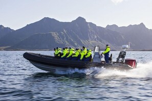Un bateau à moteur rapide transporte des passagers qui portent des gilets de sauvetage jaune vif. On aperçoit des montagnes en arrière-plan.