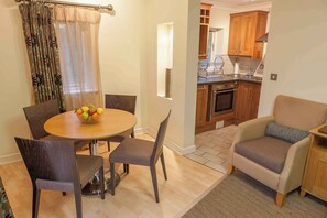 A dining area with a round table, four chairs, and a fruit bowl. A living room with a sofa and a small cabinet is visible. The kitchen is in the background with wooden cabinets and a stove.