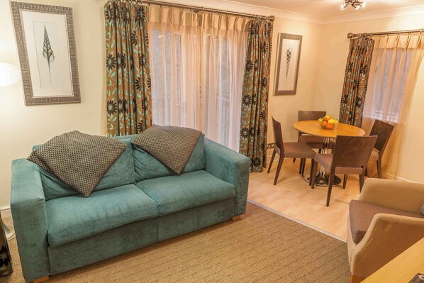 A living room with a teal sofa, patterned cushions, a dining table with chairs, and curtains with a leaf pattern.