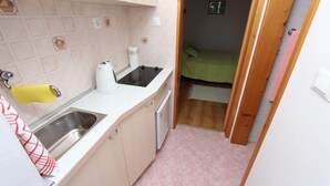 Cocina compacta con fregadero, estufa y refrigerador pequeño. Se ve una cama verde a través de una puerta abierta en la habitación contigua.