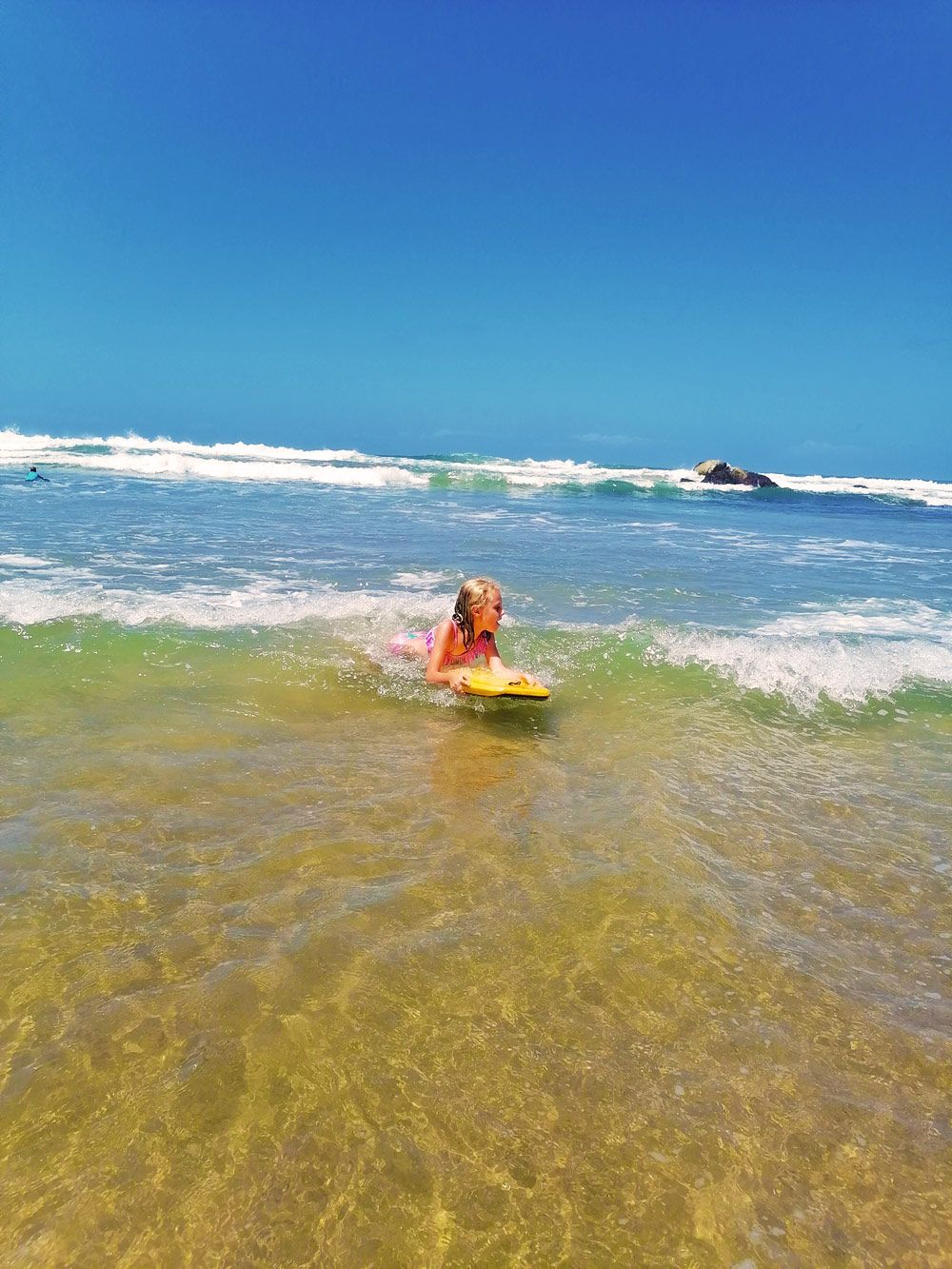 Een persoon in een zwempak op een gele surfboard in helder, ondiep water met golven, onder een strakblauwe lucht.