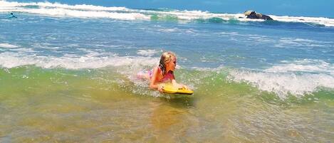 Een persoon in een zwempak op een gele surfboard in helder, ondiep water met golven, onder een strakblauwe lucht.