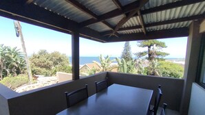 A covered outdoor patio with a table and chairs, overlooking a scenic view of the ocean and greenery. - All the Tides - Lo Tide (Margate)