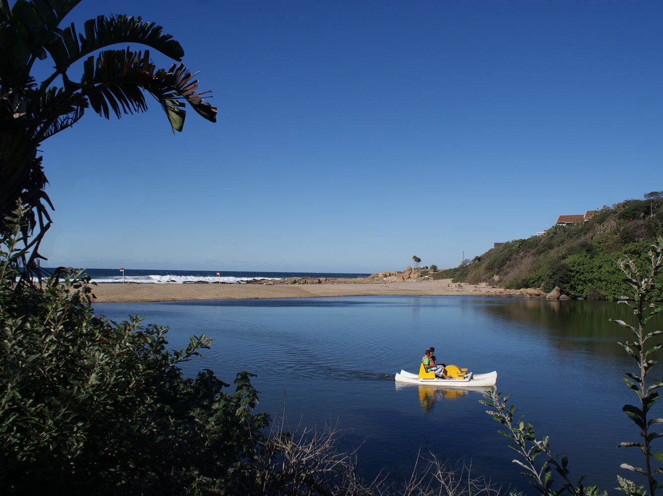 Un río tranquilo con una canoa que lleva a dos personas, rodeado de vegetación y una playa distante.