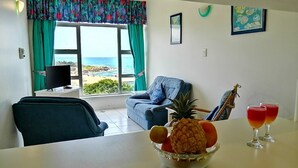 Un saló amb vistes al mar, una taula de dinar amb fruita i begudes, un sofà blau i una televisió.