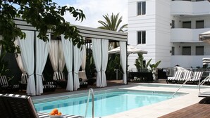 Uma área de piscina com espreguiçadeiras, um espaço de estar coberto com cortinas brancas e um edifício moderno ao fundo.