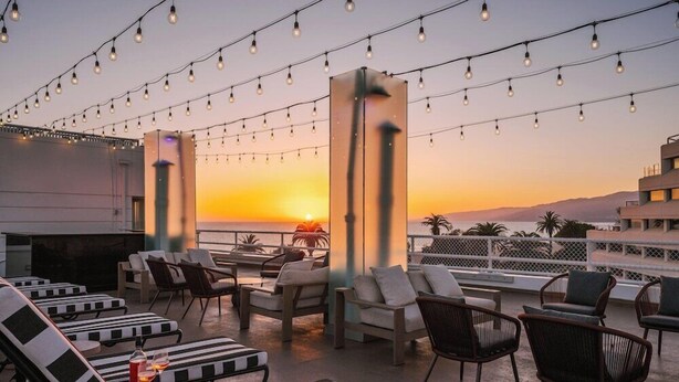 Um terraço com espreguiçadeiras e mesas, luzes de Natal e vista para o pôr do sol.