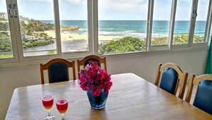 A dining table with a view of the ocean, a vase of flowers, and two glasses of red drink.