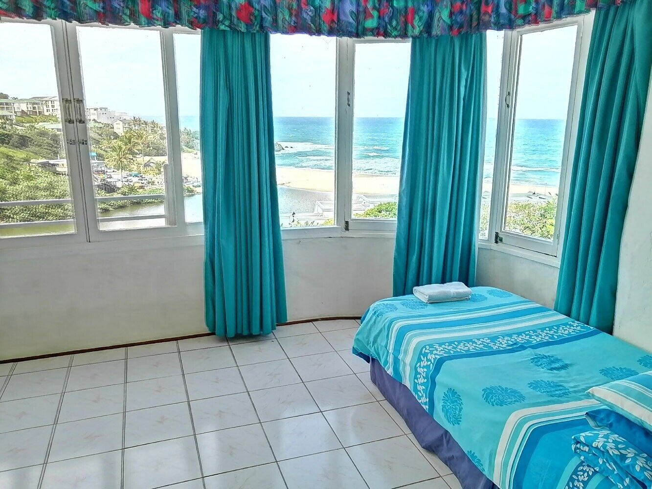 A bedroom with a bed, a window with curtains, and a view of the beach and ocean.