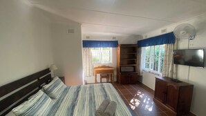 A bedroom with a large bed, a desk, a TV, and a window with curtains. - Fisherman's Cottage (Port Edward)