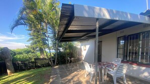 A patio with a table and chairs, a covered area, and a view of the surrounding greenery. - Fisherman's Cottage (Port Edward)