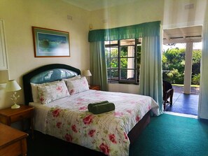 A bedroom with a bed, a nightstand, a lamp, a window with curtains, and a painting on the wall. - Greengates Cottage (Port Edward)