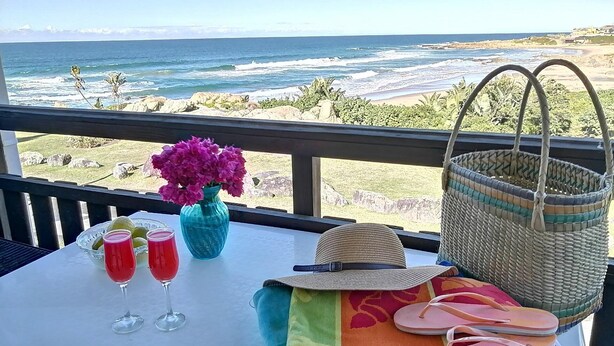 Une table avec vue sur l’océan, un vase bleu avec des fleurs roses, deux verres remplis d’une boisson rouge, un chapeau de paille, un panier tressé et une paire de tongs.