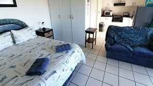 A bedroom with a bed, a nightstand, a blue sofa, a wardrobe, and a kitchenette in the background. - Rosscarbery 5 (Margate)
