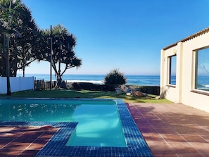 A swimming pool with a view of the ocean, surrounded by a fenced yard with palm trees and a building with large windows. - Southern Comfort - On the Pool (Margate)