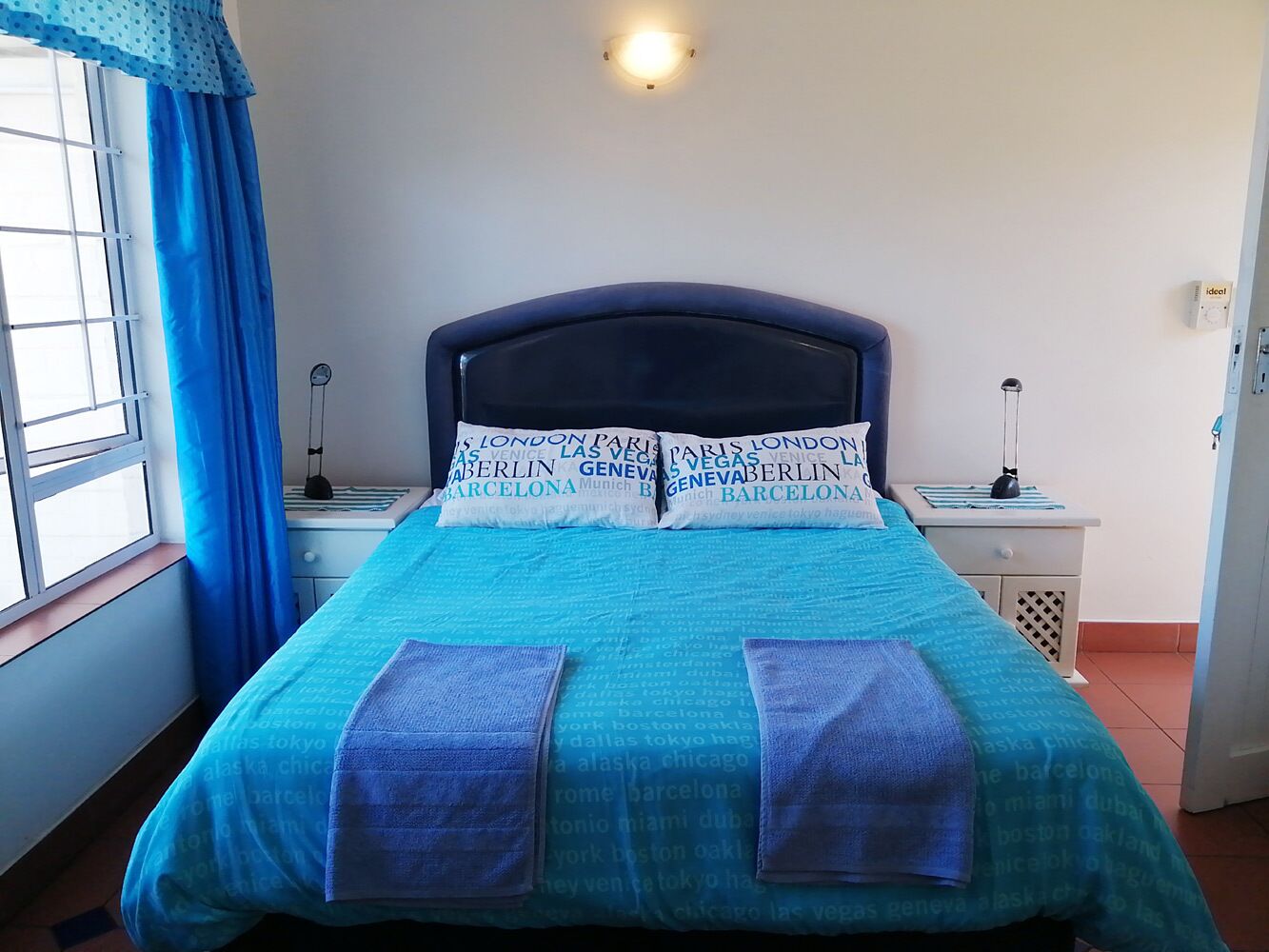 A bed with a blue bedspread and pillows, a bedside table with a lamp, and a window with blue curtains.