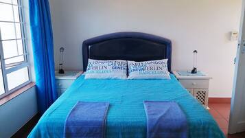 A bed with a blue bedspread and pillows, a bedside table with a lamp, and a window with blue curtains.