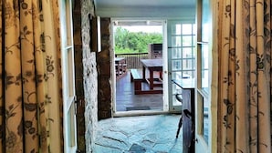 Un couloir aux murs de pierre mène à une terrasse extérieure dotée d’une table et de chaises en bois, d’un barbecue et offrant une vue sur la verdure.