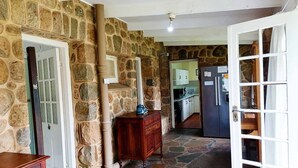 Un couloir aux murs de pierre, avec une commode en bois, une porte et une cuisine visible à travers une porte ouverte.