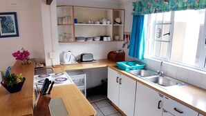 A compact kitchen with a wooden countertop, white cabinets, a window with blue curtains, and a shelf with dishes. - Southern Comfort - On the Rocks (Margate)