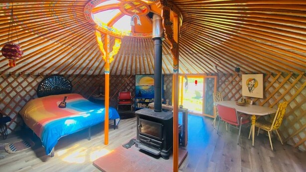 A cozy, circular yurt with a central wood-burning stove, a bed with a colorful blanket, and a small dining area.