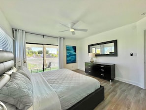 A bedroom with a bed, a dresser, a ceiling fan, a painting, and a view of the outside.