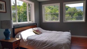 Camera da letto con un letto, un comodino, una lampada e due finestre che danno su alberi e colline.