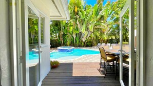 An open doorway to a pool area with a hot tub, surrounded by lush tropical plants and a wooden deck.