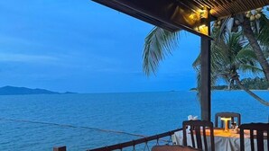 An outdoor dining area with tables and chairs, overlooking a body of water with palm trees and a wooden structure overhead. - Adarin Beach Resort (Koh Samui)