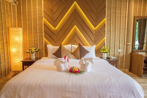 A hotel room with a large bed, wooden panel wall, and a nightstand with a lamp and flowers. - Adarin Beach Resort (Koh Samui)