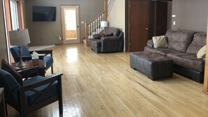 A spacious living room with a wooden floor, a large sectional sofa, a blue armchair, and a small table with a lamp. A staircase leads to an upper level. - Super Spacious Lakehome (Richmond)
