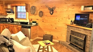 A cozy cabin interior with wooden walls, a fireplace, a kitchen area with a microwave and coffee maker, and a television displaying 'dsh Music'. - Oh Deer - Studio Wooded Twin Cabin, Hot Tub, TV & Wifi (Sevierville)