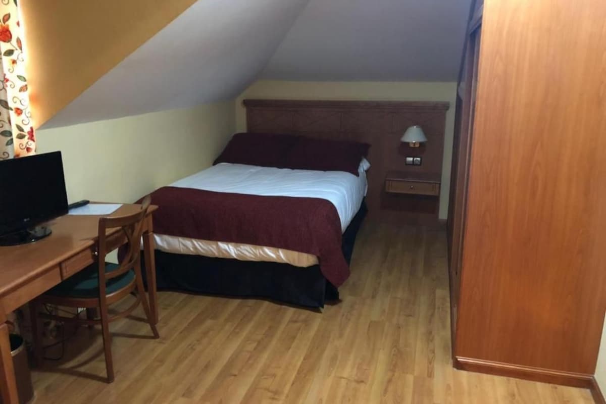 A bedroom with a bed, a desk with a computer, a chair, and a nightstand with a lamp.