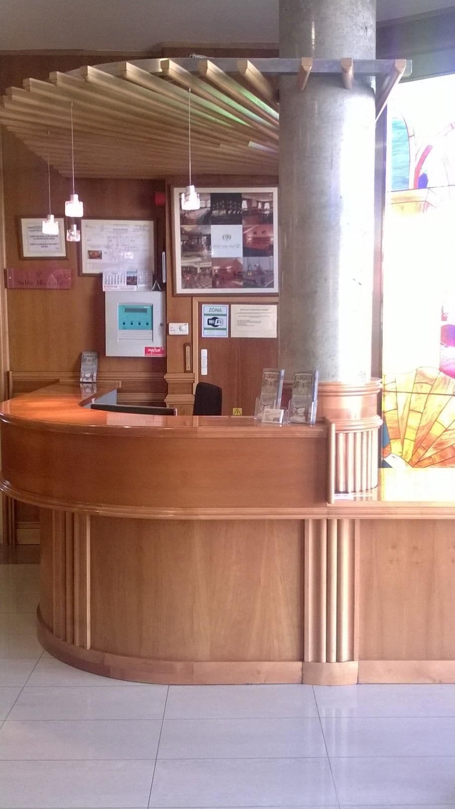 Un mostrador de recepción con detalles de madera, un pilar grande, lámparas colgantes y varios carteles informativos en la pared.