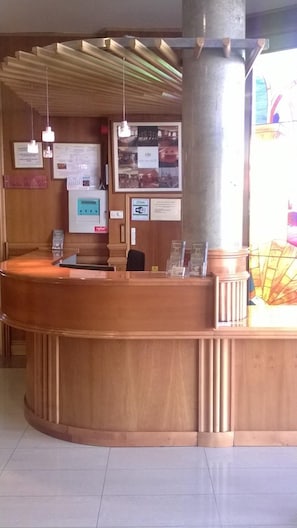 A reception desk with a wooden finish, a large pillar, hanging pendant lights, and various informational posters on the wall. - Hotel Camino Real (Valdefresno)