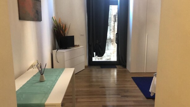 Une pièce avec un plancher en bois, un bureau orné d’un vase contenant des roseaux, une chaise, une petite table recouverte d’une nappe verte, et une vue sur un bâtiment à travers une fenêtre munie de rideaux sombres.