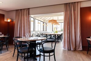Een restaurant met ronde tafels, zwarte stoelen en een groot raam met gordijnen.