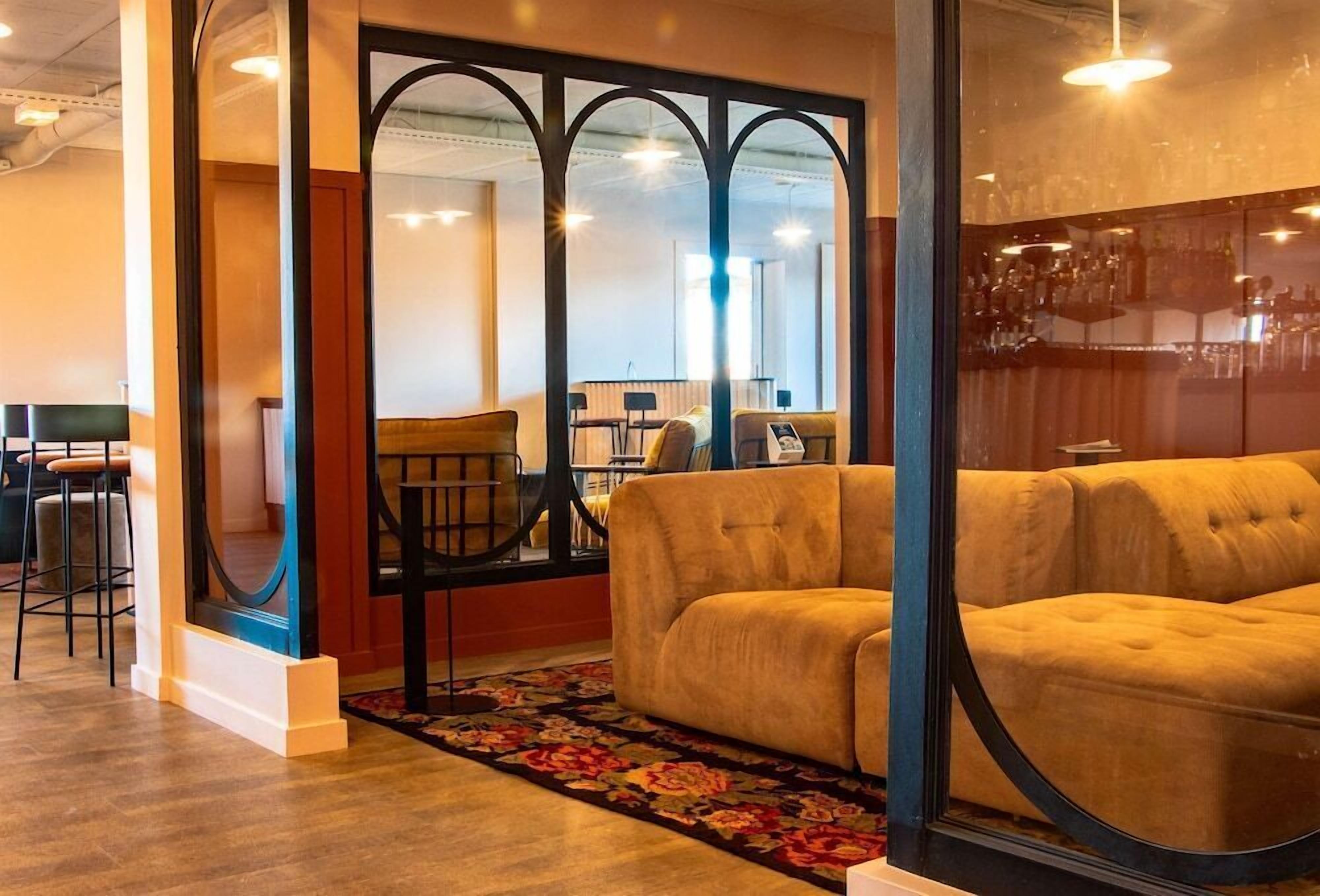 Un espace salon confortable avec un canapé et des fauteuils, un tapis à motifs et une cloison en verre donnant sur un bar.